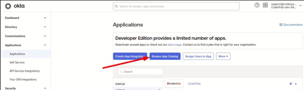 The image shows a screenshot of the Okta dashboard, specifically under the "Applications" section. A red arrow points to the "Browse App Catalog" button, which is next to the "Create App Integration" button. The left sidebar menu includes options like Dashboard, Directory, Customizations, and Applications, among others. The top-right corner displays a support email and the user account details.